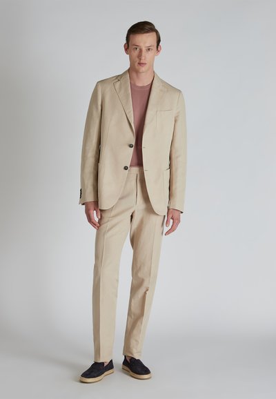 Abito di lino beige con una tasca sul petto, bottoni neri e pantaloni coordinati. Indossato sopra una camicia rosa chiaro e espadrillas nere.