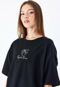 Camiseta negra de gran tamaño hecha de algodón, con un corazón bordado en blanco que presenta un diseño de globo y el texto "Project Paris" debajo.
