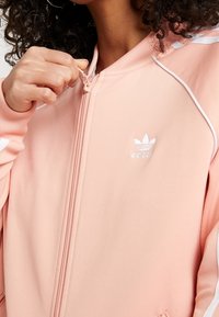 Světle růžová bunda na zip s bílými doplňky, vyrobená z hladké látky. Na levé straně hrudi je malé bílé logo Adidas a kontrastní lemování.