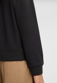 Zwarte, textuur-sweatshirt mouw met ribbelmanchet, gecombineerd met beige stoffen broek. Focus op materiaaleigenschappen en breipatronen.