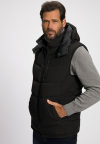 Schwarze Steppweste mit abnehmbarer Kapuze, ausgestattet mit einem Reißverschluss vorne, seitlichen Taschen und einem gesteppten Design. Graues Langarmshirt darunter.
