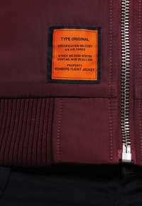 Giacca bomber bordeaux con polsini a coste, chiusura frontale con zip e una etichetta arancione che dettaglia specifiche e proprietà.