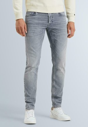Muž nosiaci vyblednuté sivé slim-fit džínsy a biele tenisky stojaci pred hladkým svetlým pozadím.