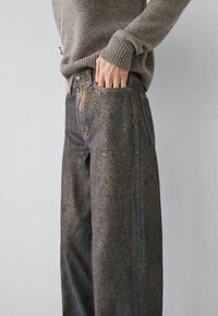 Jean bleu foncé avec un éclat métallique texturé. Coupe ample, design classique à cinq poches, associé à un pull en tricot gris.