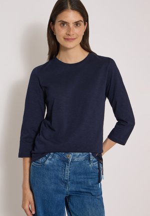 Frau mit langen braunen Haaren, die ein marineblaues Dreiviertelarm-Top und blaue Jeans trägt, steht mit einer Hand in der Hüfte.