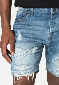 Τζιν σορτσάκια σε ανοιχτό μπλε με φθαρμένες λεπτομέρειες, ακατέργαστο στρίφωμα και εκτεθειμένο εσωτερικό ύφασμα. Διαθέτουν πέντε τσέπες και κούμπωμα με κουμπί.