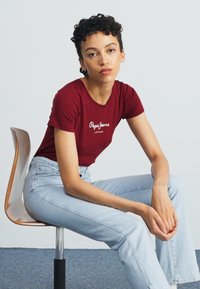 Burgundinė prigludusi marškinėliai su "Pepe Jeans" logotipu, dėvimi su šviesiai mėlynomis plačiomis džinsinėmis kelnėmis. Asmuo sėdi ant medinės kėdės prieš paprastą fono.