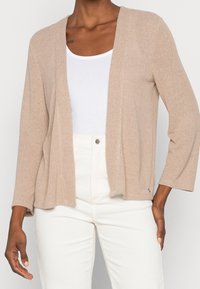 Person klädd i en beige öppen cardigan över en vit topp och vita byxor med hög midja, stående mot en enfärgad bakgrund.