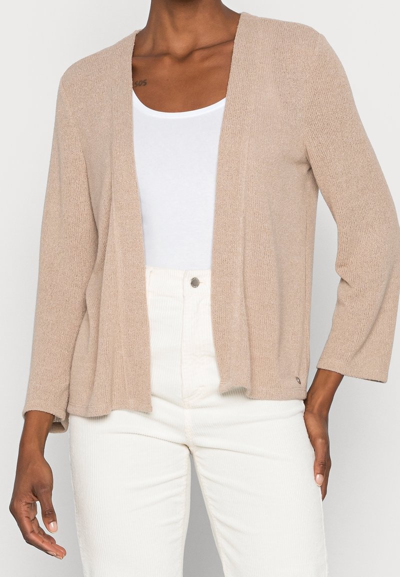 Person klädd i en beige öppen cardigan över en vit topp och vita byxor med hög midja, stående mot en enfärgad bakgrund.