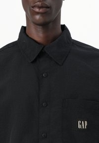Camisa negra de botonadura clásica con cuello, tejido texturizado, bolsillo en el lado izquierdo del pecho y logotipo 'GAP' en blanco en el bolsillo.