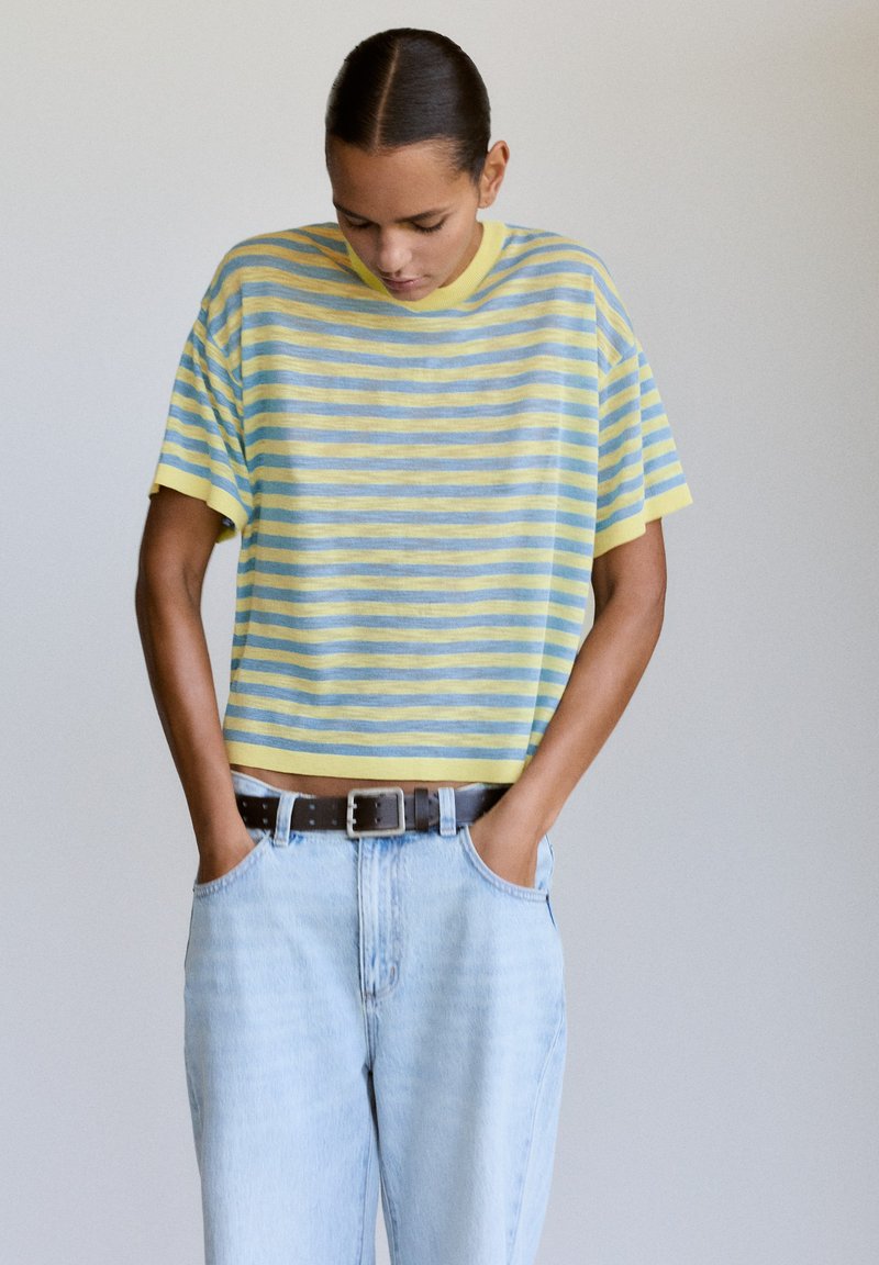 Personne portant une chemise à manches courtes rayée jaune et bleu et un jean bleu clair avec une ceinture noire, regardant vers le bas les mains dans les poches.