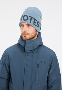 Bonnet bleu avec "PROTEST" en lettres foncées, associé à une veste zippée bleu marine avec un col haut et une poche zippée.