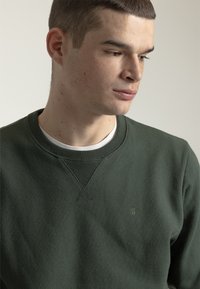Sudadera de color verde oscuro hecha de tela texturizada. Cuenta con un cuello redondo, puños acanalados y un pequeño logo en el pecho.