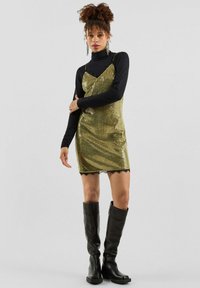 Frau in schwarzem Rollkragenpullover unter einem goldenen Pailletten-Kleid, kombiniert mit schwarzen kniehohen Stiefeln und hängenden Ohrringen vor grauem Hintergrund.
