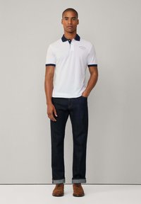Bílá polo košile s námořnickým límečkem a manžetami, spárovaná s tmavými denimovými džíny. Hnědé boty dokončují vzhled. Minimalistický design s detailem loga.