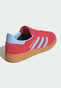 Červené suède tenisky Adidas Spezial s modrými pruhmi, žltým logom a gumovou podrážkou, zobrazené zo zadnej strany na bielom pozadí.