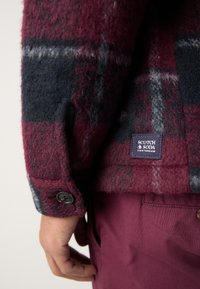 Veste en laine à carreaux bourgogne et marine avec des détails en boutons noirs et une étiquette en tissu. Présente une texture douce et un design structuré.