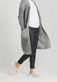 Lång, texturerad cardigan i svart och vitt över en vit blus, kombinerad med svarta leggings och ljusrosa sneakers med vita sulor.