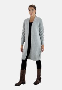 Helles graues Strickcardigan mit gerippter Textur, offener Front und Pufferärmeln. Kombiniert mit schwarzen Leggings und braunen kniehohen Stiefeln.