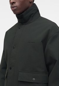 Άνδρας που φοράει μαύρο μπουφάν Carhartt με ψηλό γιακά, κουμπωτό εμπρός και μεγάλες τσέπες με καπάκι, σε απλό ανοιχτό φόντο.