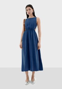 Vestido azul sin mangas con cintura ajustada, falda larga, hecho de una tela suave. Combinado con unas flats plateadas y un bolso de hombro.