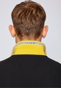 Homme aux cheveux bruns courts vu de dos portant une chemise noire avec un col jaune montrant un texte répété à l'intérieur.