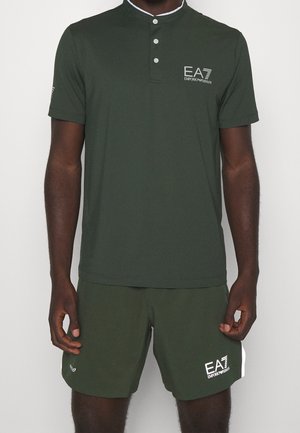 Mand iført mørkegrøn kortærmet polo og matchende shorts med hvide EA7 Emporio Armani logoer på bryst og ben.