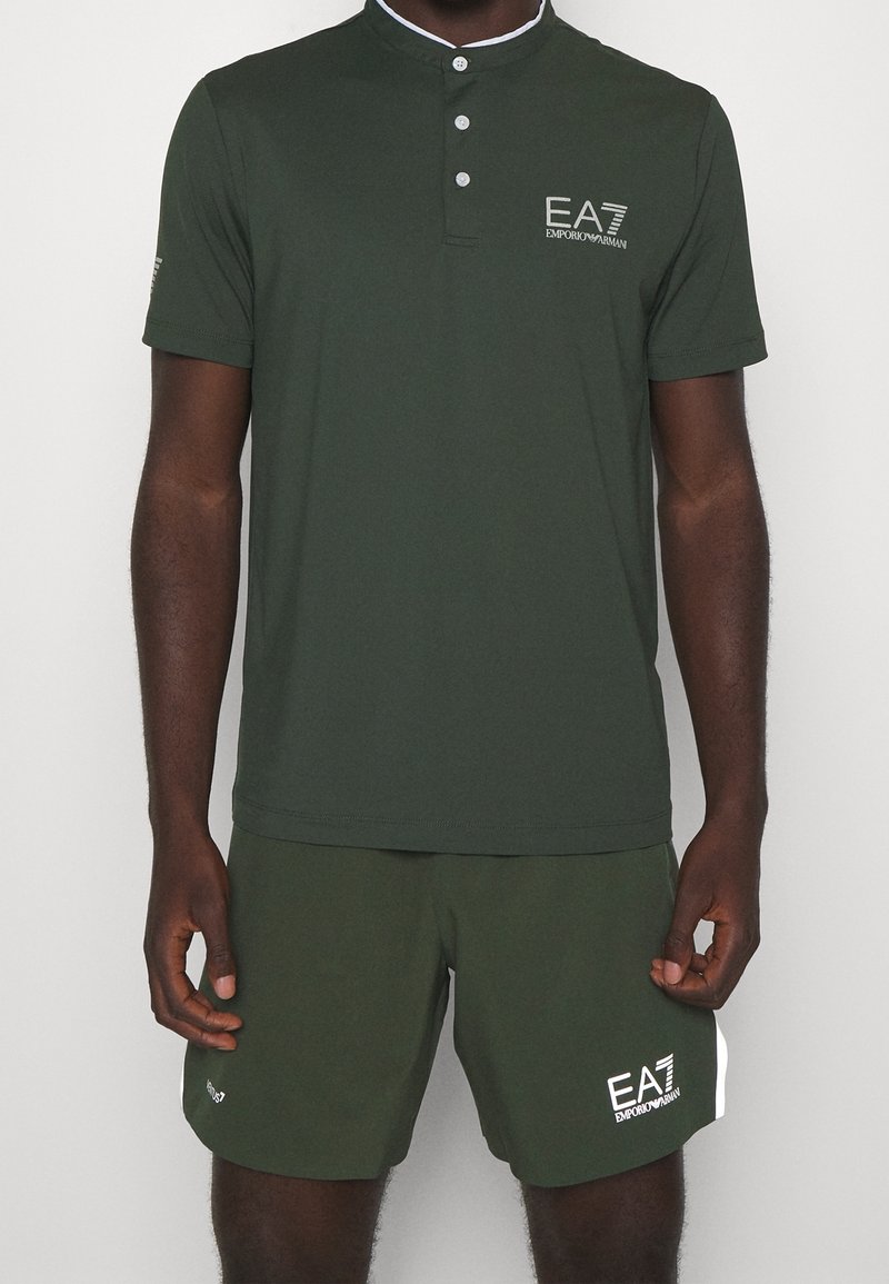 Homme portant un polo à manches courtes vert foncé et un short assorti avec des logos blancs EA7 Emporio Armani sur la poitrine et la jambe.
