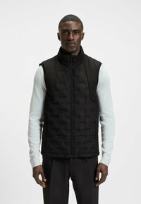 Svart vadderad väst med hög krage, dragkedjestängning och fickor, som bärs över en ljusblå långärmad skjorta med en slät textur.