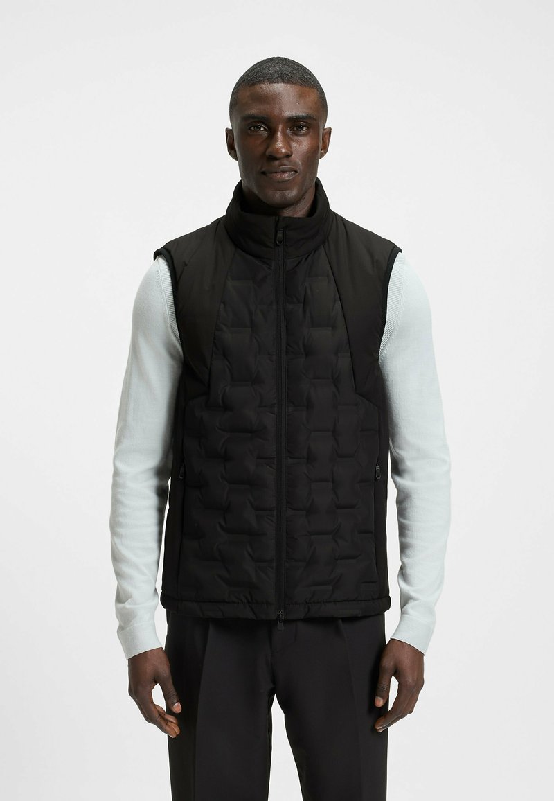 Svart vadderad väst med hög krage, dragkedjestängning och fickor, som bärs över en ljusblå långärmad skjorta med en slät textur.