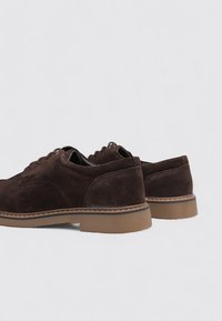 Zapatos de ante marrón con punta redonda, suela de goma marrón claro y costuras en contraste. Presentan un diseño clásico con cordones.