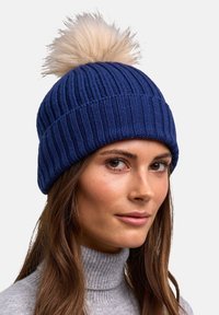 Vrouw met lang bruin haar die een blauwe gebreide beanie draagt met een grote beige pompon en een lichtgrijze coltrui.
