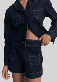 Ensemble en denim foncé avec une veste et un short, présentant des coutures claires, avec deux poches à l'avant de la veste et des poches assorties sur le short.
