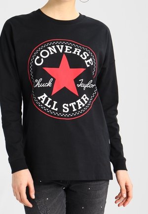 Person trägt ein schwarzes Langarmshirt mit einem großen roten Stern und dem weißen "Converse All Star Chuck Taylor"-Logo auf der Vorderseite.