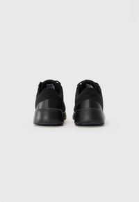 Par de zapatillas deportivas negras mostradas desde la parte trasera sobre un fondo claro y liso, destacando suelas gruesas y cuellos acolchados.