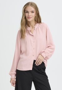 Light pink blouse with a ruffled neckline and cuffs, featuring a button placket and soft fabric, paired with black pants.