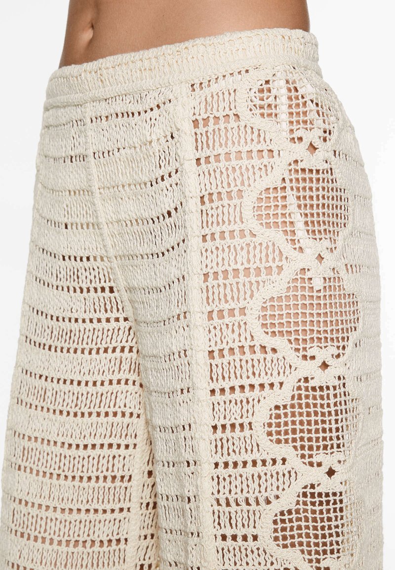 Gros plan sur une personne portant un pantalon en crochet couleur crème avec des motifs géométriques en ajouré sur le côté, laissant entrevoir la peau en dessous.