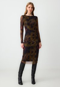 Langdachtige, getailleerde jurk in donkerbruin en zwart met een tie-dye patroon. Heeft een verzamelde taille en een lengte tot halverwege de kuit. Gecombineerd met laarzen tot de knie.
