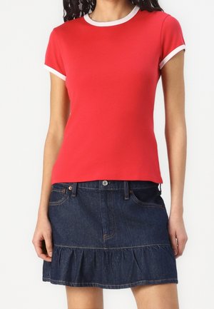 Vrouw draagt een nauwsluitend rood-shirt met korte mouwen met witte bies en een donkerblauwe denim rok met een gerafelde zoom.