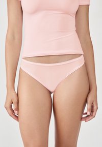 Frau in hellrosa nahtloser Unterwäsche und passendem figurbetontem Kurzarmoberteil, steht mit entspannten Armen an den Seiten vor einem weißen Hintergrund.