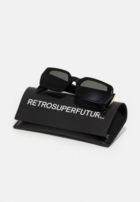 Mustad ristkülikukujulised prillid tumedate linsidega mustal pehmest kotil. Kotil on valgete tähtedega kiri "RETROSUPERFUTURE".