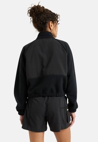 Čierna bunda s fleece textúrou na hornej polovici, kontrastujúca s hladkým materiálom nižšie. Má elastický pás a dlhé rukávy.