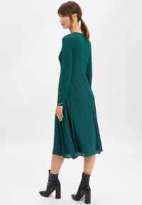 Kobieta o brązowych włosach związanych w kucyk, ubrana w długą, plisowaną zieloną sukienkę midi z długim rękawem oraz czarne botki na obcasie, stojąca bokiem.