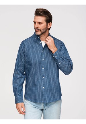 Mand justerer kraven på en blå skjorte med lange ærmer og små hvide mønstre, iført lyseblå jeans, neutral baggrund.