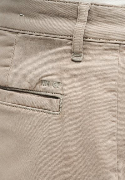 Gros plan sur un tissu beige montrant une poche arrière cousue et une boucle de ceinture sur un pantalon avec une broderie discrète « NW. Q7 ».