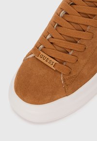 Brown suede sneaker with flat laces, rubber sole, and a beige logo tag on the side. Textured upper and smooth toe cap.