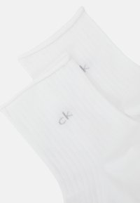 Chaussettes blanches côtelées avec une texture douce, dotées d'un petit logo "ck" gris sur le côté. Le design met l'accent sur le confort et un style simple.