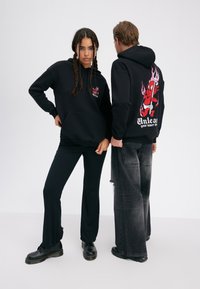 To personer iført sorte hættetrøjer med røde djævelgrafikker; den ene vender front mod kameraet med hænderne på hofterne, den anden viser rygdesignet og holder en trefork.