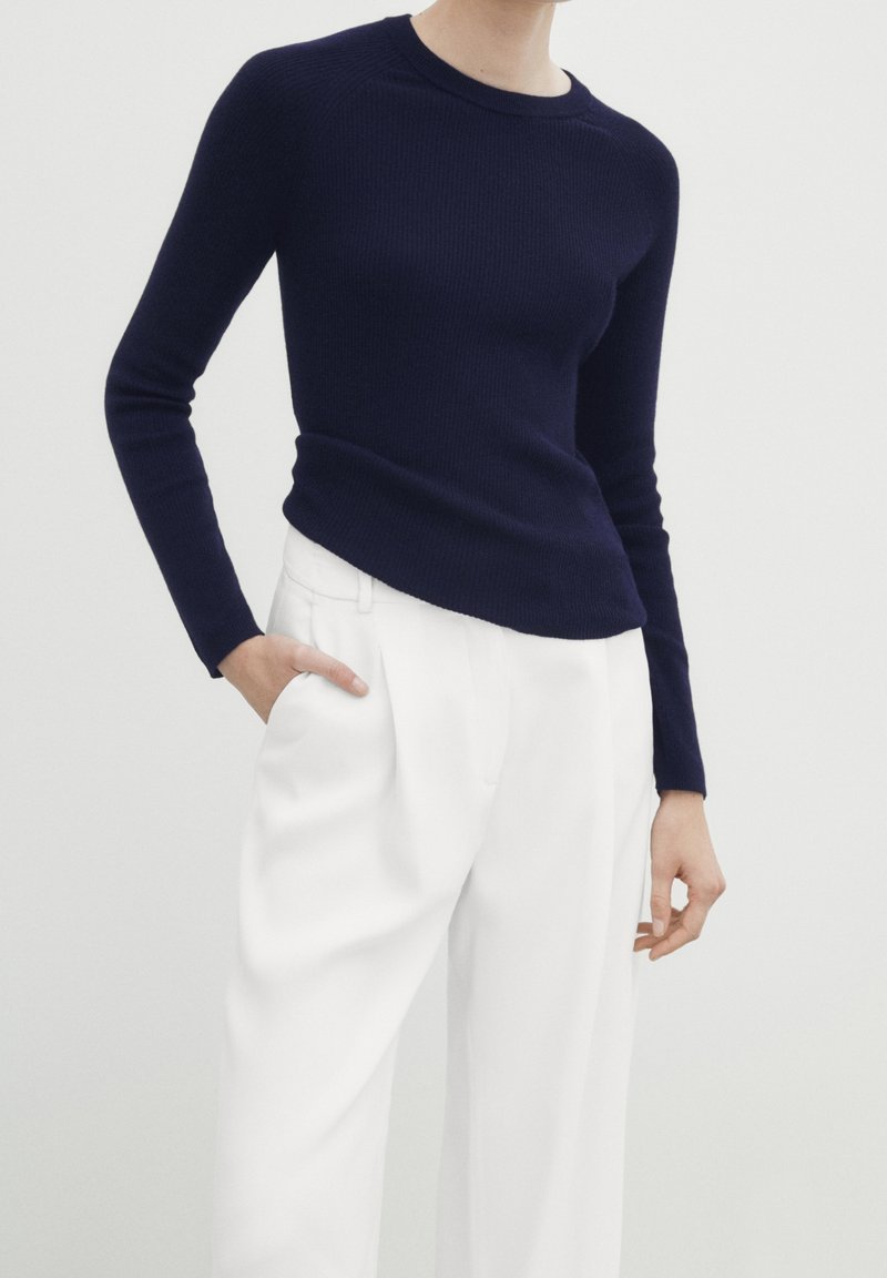 Personne portant un pull ajusté à manches longues bleu marine et un pantalon blanc taille haute à jambes larges, une main dans la poche.