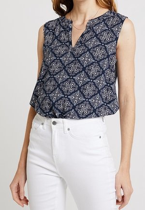 Vrouw draagt een mouwloze donkerblauwe top met witte bloemmotieven en witte hoge taillebroeken, staand met ontspannen armen langs haar zij.
